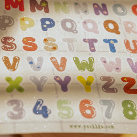 A close-up of a sticker sheet featuring colorful, glittery bubble-style alphabet letters and numbers from M to 8 on a white background.
