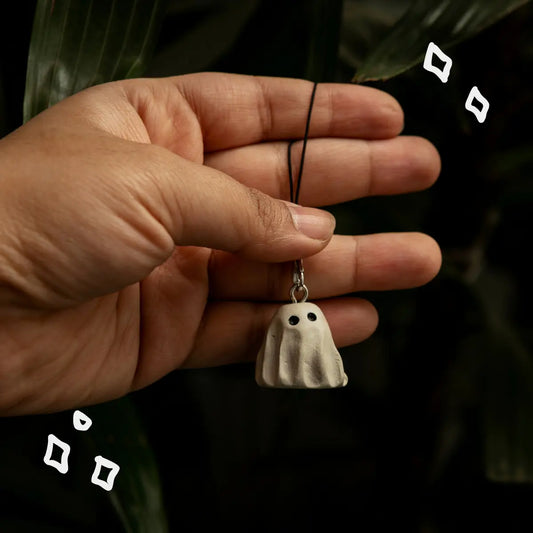 A hand holding a small ghost-shaped kaychain with a black cord and silver clasp, set against a dark background with blurred green leaves.