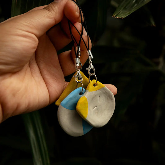 A hand holds three small clay accessory pieces with black cords and silver clasps against a dark, leafy background. The pieces are mostly light gray with yellow and blue painted sections.
