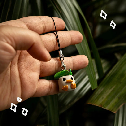 A hand holding a small duck-shaped charm on a black cord, with dark green leaves in the background. The charm is white with orange feet and a green hat, and has small black eyes.