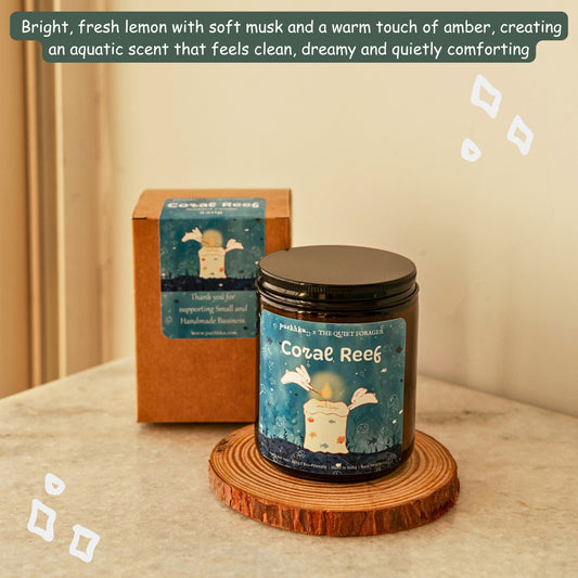 A candle jar labeled 'Coral Reef' sits on a round wood coaster in front of its matching product box. The scene is styled on a tabletop against a warm beige wall, with a scent description banner at the top describing a bright lemon, soft musk, and amber aquatic scent.