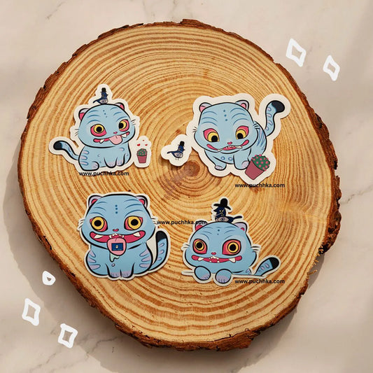 Four cartoon cat stickers in light blue with colorful expressions are arranged on a round wood slice background. The stickers have white borders and small printed website text, photographed on a light marble-like surface.