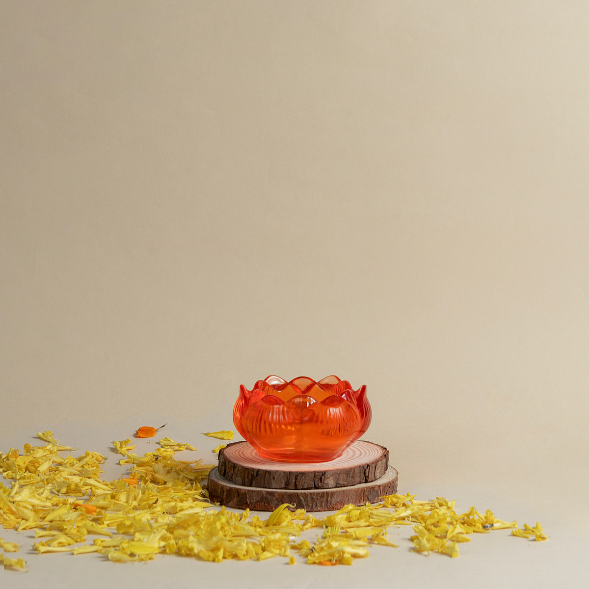 A translucent orange-red lotus-shaped bowl or candle holder displayed on two stacked wood slices, with scattered yellow flower petals on a beige surface against a plain beige background.