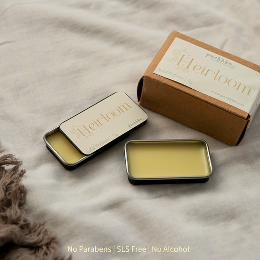 A small rectangular metal tin of solid perfume balm next to an open tin showing the pale yellow perfume balm, with a kraft paper box labeled Heirloom . The products are arranged on a textured beige fabric background.