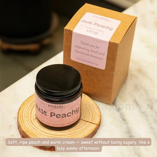 A small jar of Just Peachy scented candle with a black lid and pink label sits on a round wood slice coaster, with its kraft paper box behind it on a marble surface. The brand name 'Puchhka' is visible on both the jar label and the box.