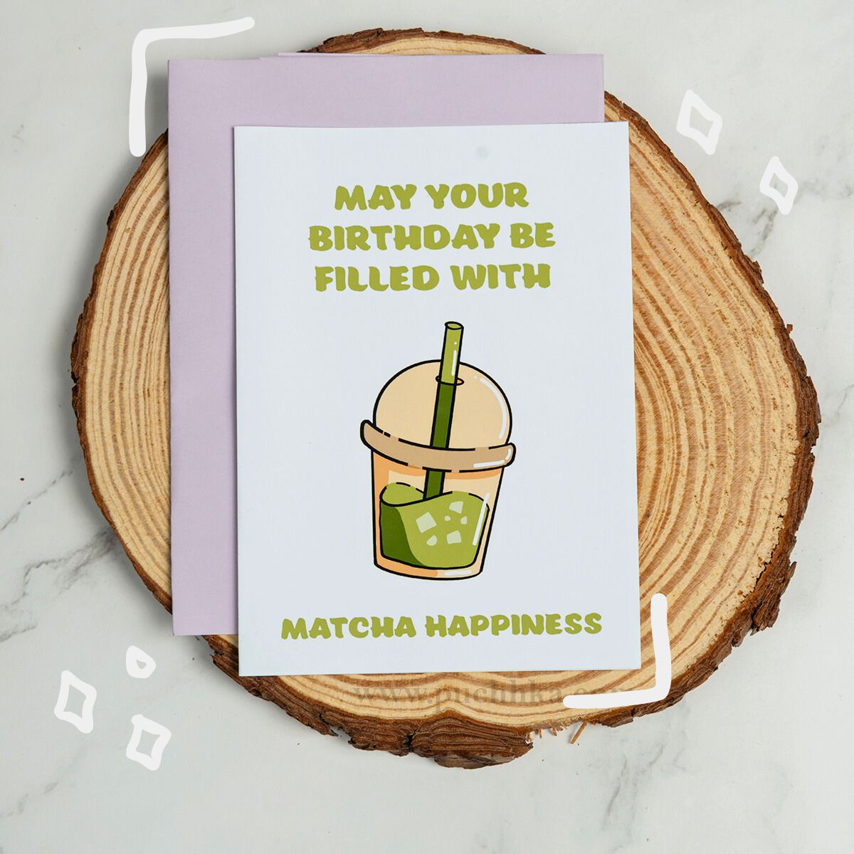 A square birthday card with a light purple envelope, placed on a round wood slice over a white marble surface. The card has a white front with green text reading 'MAY YOUR BIRTHDAY BE FILLED WITH MATCHA HAPPINESS' and an illustration of a matcha drink with a bamboo straw.