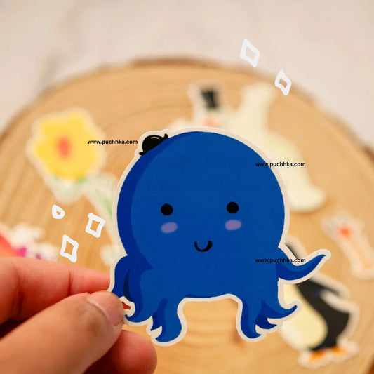 A hand holds a die-cut sticker of a cute blue octopus with a small black hat, set against a blurred background of other stickers on a wooden surface.