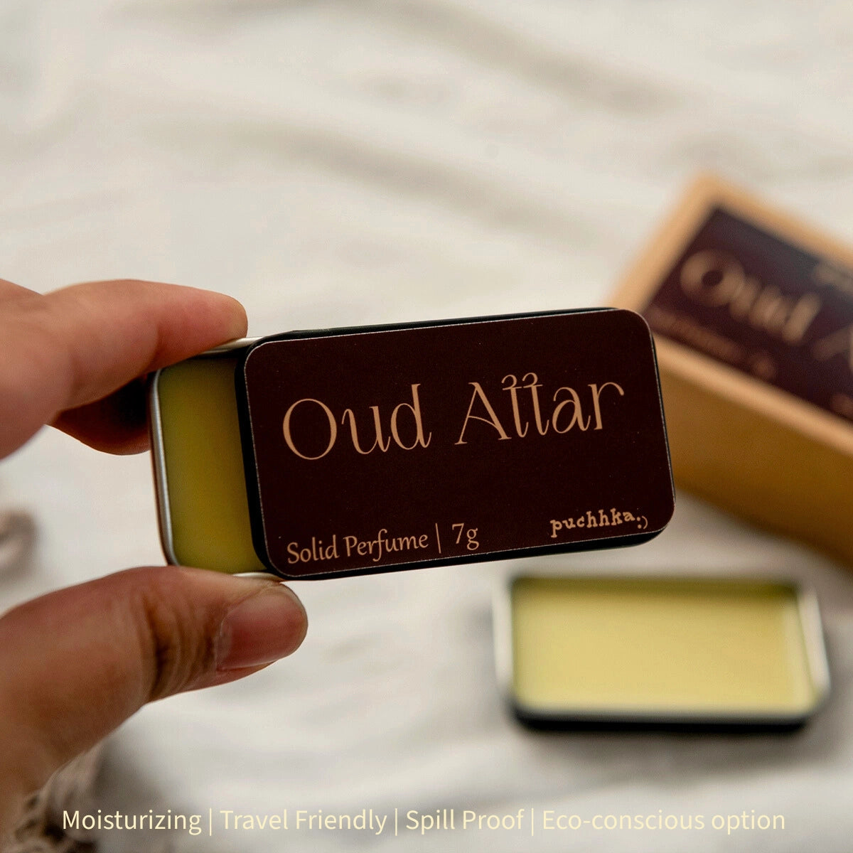 A hand holds a small rectangular metal tin of solid perfume labeled 'Oud Attar' against a softly blurred light background. Another open tin and a branded box are blurred in the background.