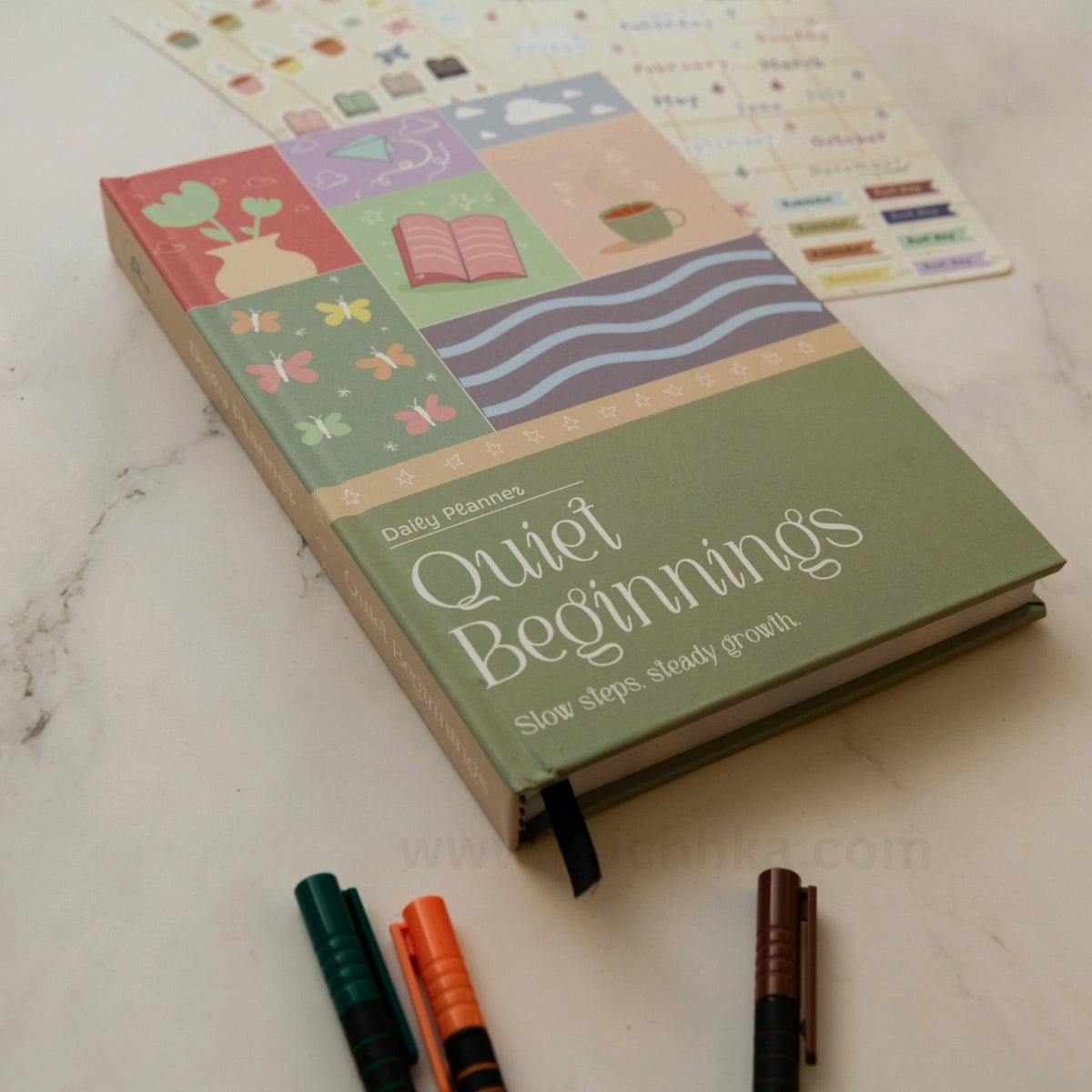 A hardcover daily planner titled 'Quiet Beginnings' lies on a light marble surface, with sticker sheets and three colored pens nearby. The cover is muted sage green with pastel illustrated panels and white script text.