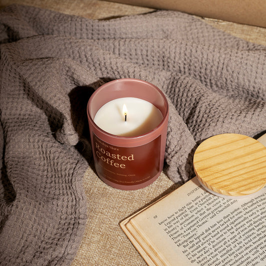 A lit candle in a dusty rose glass jar labeled 'Roasted Coffee' by Daakshya Store (now Puchhka), styled on a beige fabric surface with a gray waffle-knit blanket, an open book, and a round wooden lid nearby.