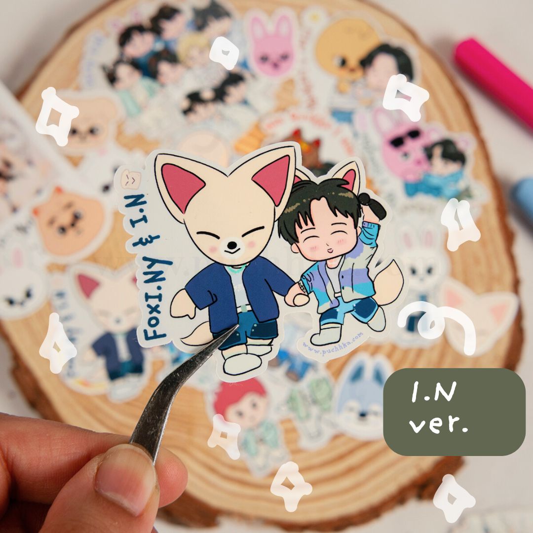 A hand holds a die-cut sticker featuring two cartoon characters, FoxiNY and IN, with a small metal tweezler supporting the sticker, set against a background of many other colorful stickers on a wood slice.
