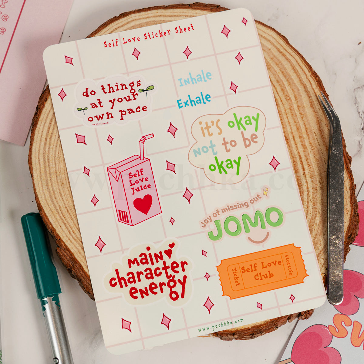 A self-love sticker sheet displayed on a rustic wood coaster, surrounded by a green pen on a marble surface. The sticker sheet features various colorful retro-style stickers with positive affirmations like 'Main character energy' and 'it's okay not to be okay'.