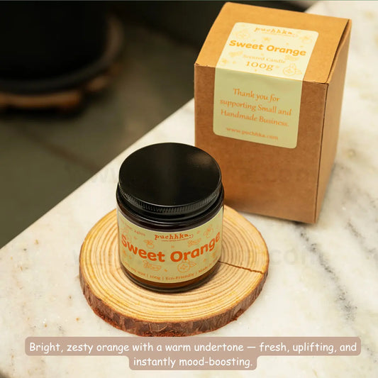 A jar of Puchhka Sweet Orange scented candle placed on a round wooden coaster, with its kraft paper box standing behind it on a white marble surface.