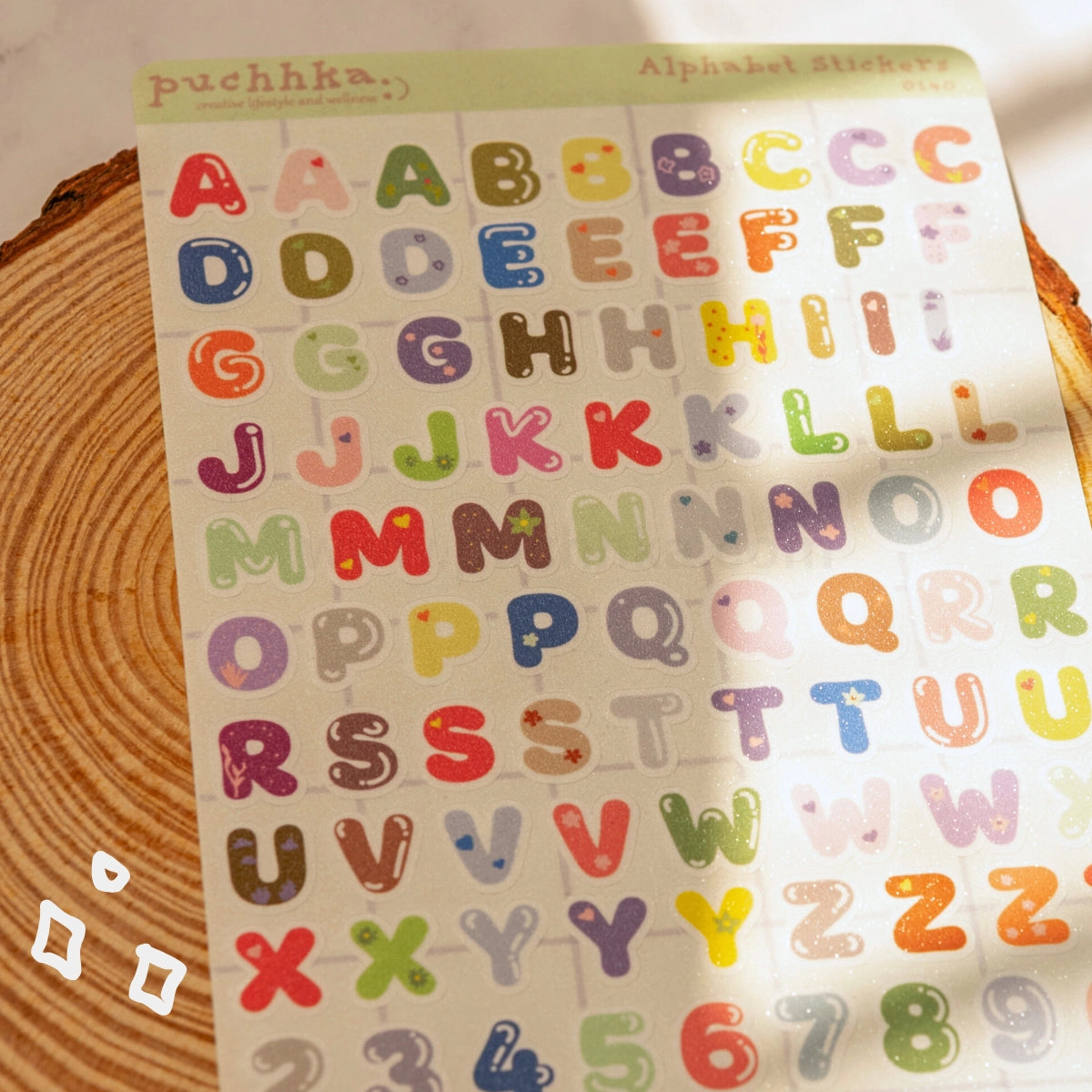 A sheet of colorful alphabet stickers displayed on a wooden surface. The stickers feature uppercase letters with cute illustrations like hearts, stars, flowers, and dots in various pastel and bright colors.