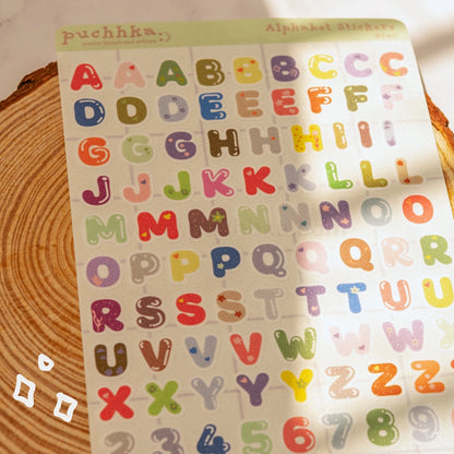 A sheet of colorful alphabet stickers displayed on a wooden surface. The stickers feature uppercase letters with cute illustrations like hearts, stars, flowers, and dots in various pastel and bright colors.