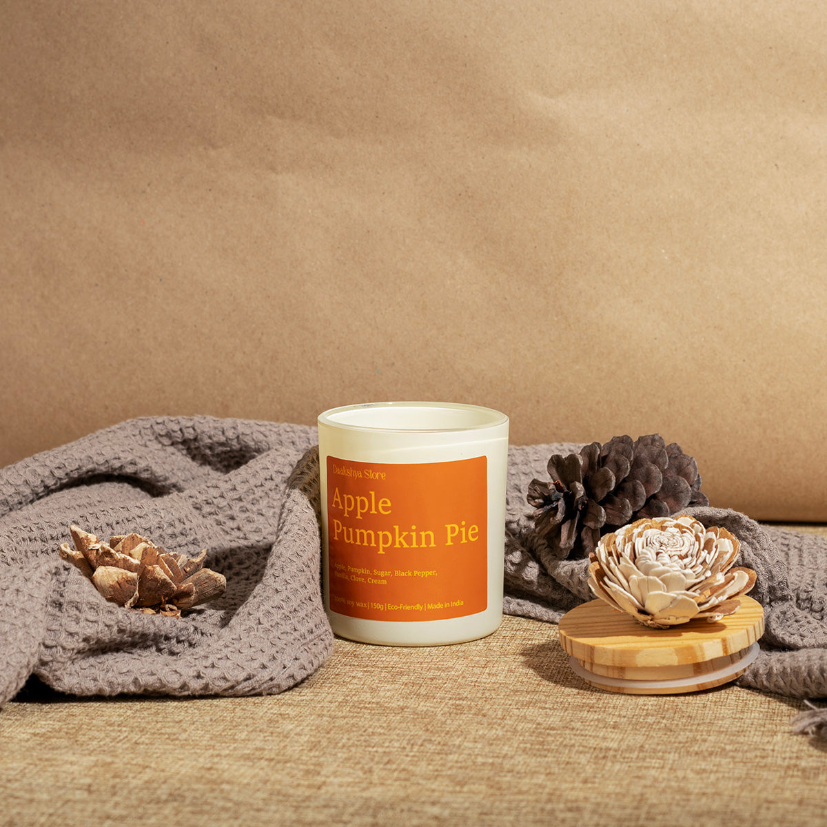 A candle in a white jar with an orange label reading 'Apple Pumpkin Pie' is styled on a burlap surface with a gray knit blanket, pinecones, and a dried flower against a brown paper backdrop.
