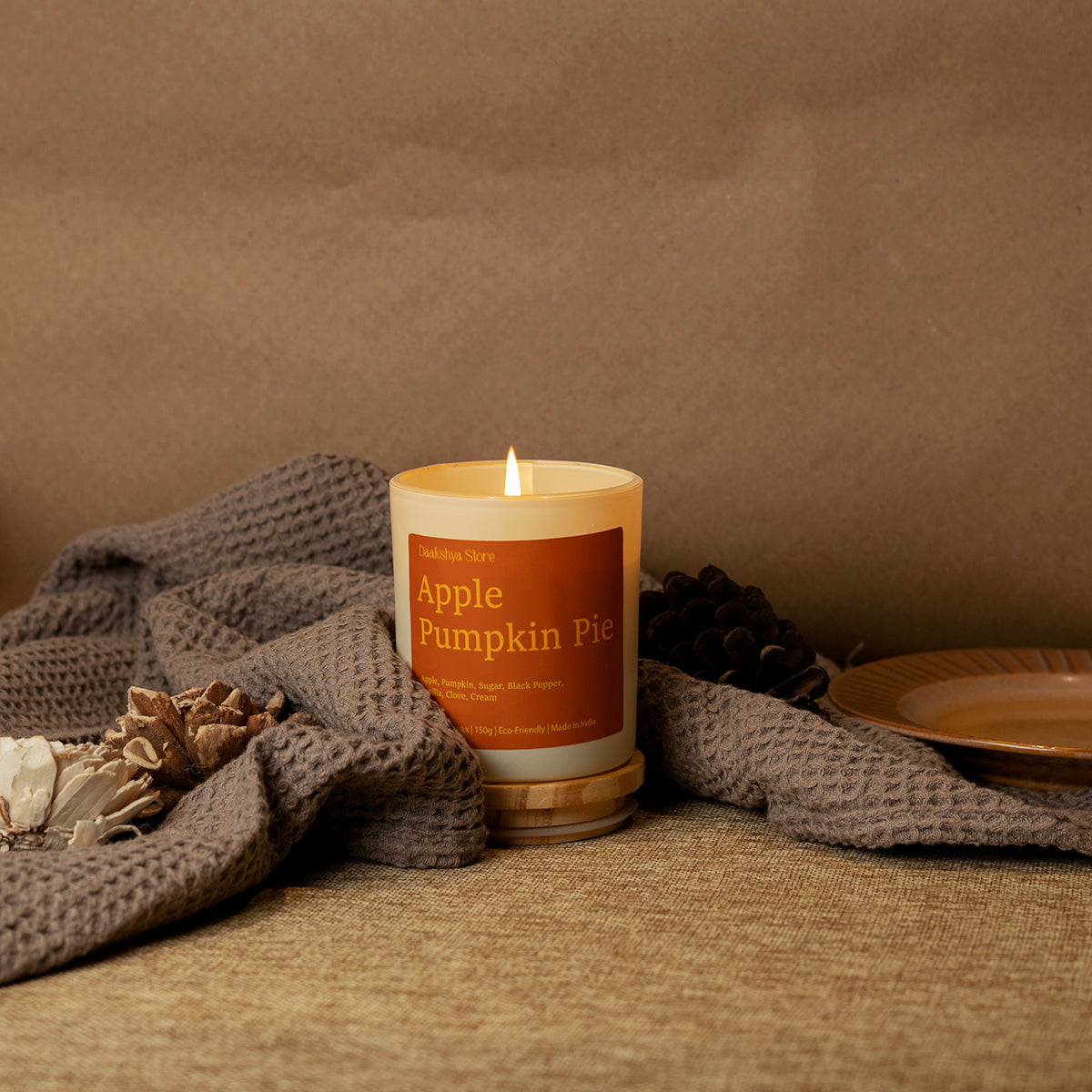 A lit candle in a cream-colored glass jar with a warm orange label reading 'Apple Pumpkin Pie' by the brand 'Puchhka', styled on a textured beige surface with a gray knit blanket, pinecones, and a ceramic plate against a brown backdrop.