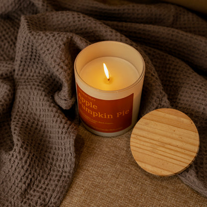 A lit candle in a frosted glass jar with a warm orange label reading 'Apple Pumpkin Pie,' placed on a textured brown blanket with a round wooden lid beside it.