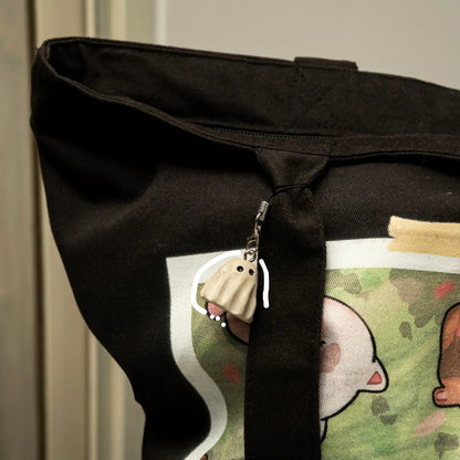 Close-up of a black canvas tote bag with a printed cartoon graphic on the front and a small ghost-shaped keychainl, photographed indoors against a neutral background.