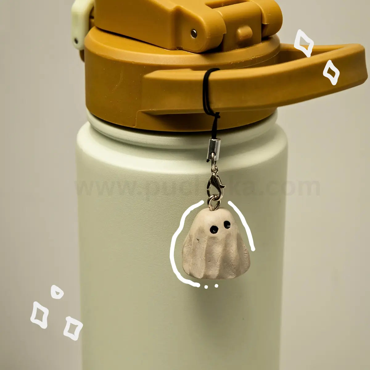 A close-up of a white insulated water bottle with a mustard yellow lid, featuring a small ghost-shaped charm hanging from the strap.