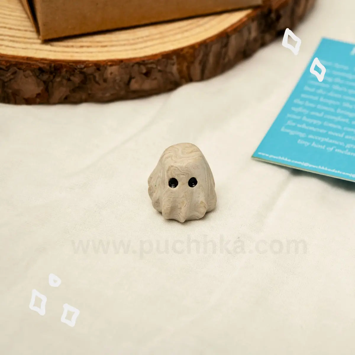 A small beige clay desk buddy with black eyes sits on a white fabric surface, with a wood slice, a blue card, and a brown box partially visible in the background.