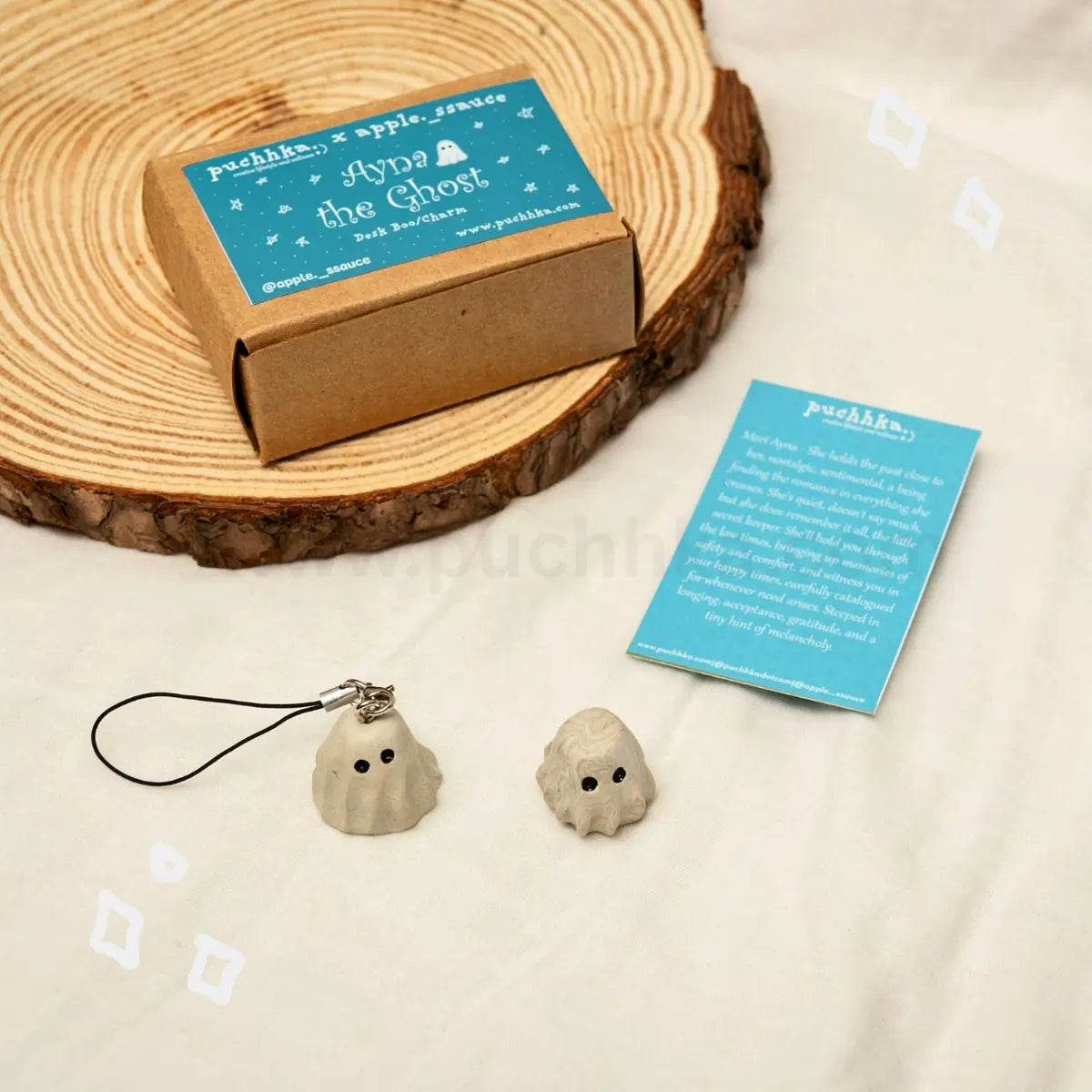 Two small ghost-shaped keychains in a light beige clay material with black strings, displayed on a white fabric surface next to a brown kraft box, a blue product card, and a round wood prop. The brand name 'puchhka' is visible on the packaging and card.