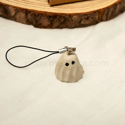 A small ghost-shaped charm in a light warm gray tone with two black eyes and a black cord loop, photographed on a light surface with a wood slice in the background.