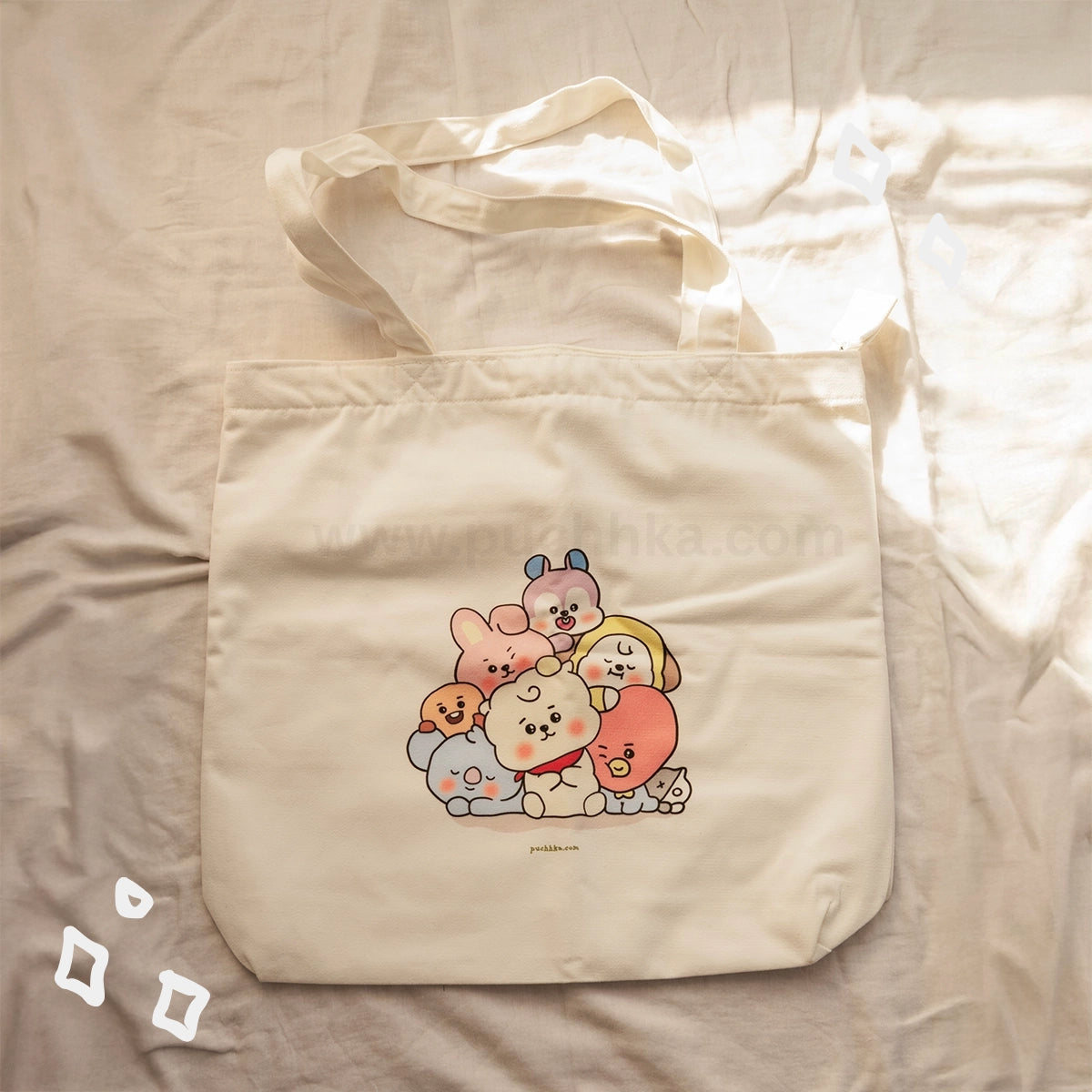 A cream-colored canvas tote bag featuring a cute group illustration of BTS characters, laid flat on a beige fabric background with warm sunlight filtering in from the right side.