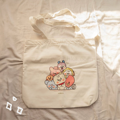 A cream-colored canvas tote bag featuring a cute group illustration of BTS characters, laid flat on a beige fabric background with warm sunlight filtering in from the right side.