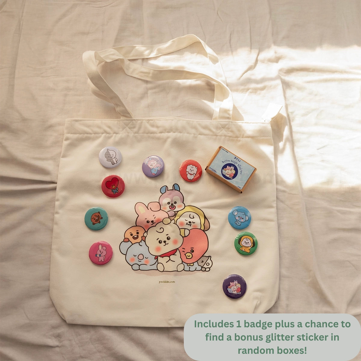 A cream canvas tote bag laid flat on a light fabric surface, decorated with a colorful group of cute BT21 characters and surrounded by several round pin badges and a small sticker box. A text bubble at the bottom right says it includes 1 badge plus a chance to find a bonus glitter sticker in random boxes.