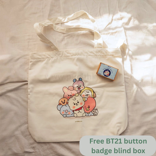 A cream-colored canvas tote bag featuring a colorful group illustration of cute cartoon characters, laid flat on a beige fabric background. A small blue and orange box labeled BT21 is attached to the bag, and a green text overlay at the bottom right reads 'Free BT21 button badge blind box'.