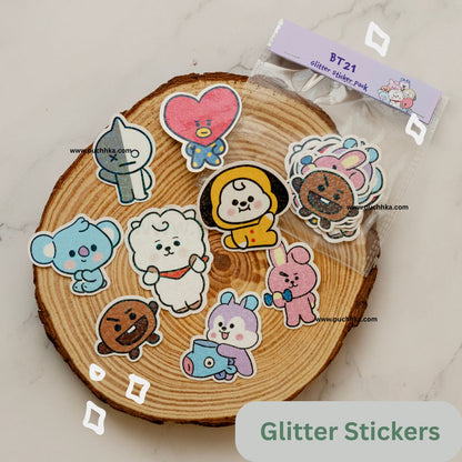 A set of BT21 glitter stickers displayed on a round wood  coaster, with the clear plastic packaging bag visible at the top right. The stickers feature various cute BT21 characters in colorful glitter finishes.