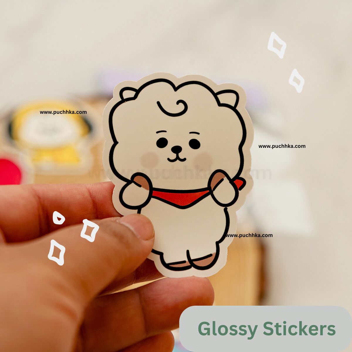 A hand holds a glossy die-cut sticker of a cute white fluffy character with a red collar against a softly blurred indoor background. 