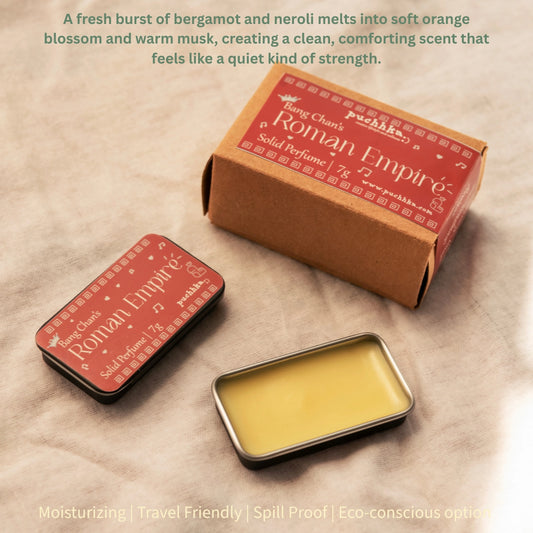 A small rectangular metal tin of solid perfume with pale yellow perfume sits in front of its brown cardboard box and a matching closed tin, all labeled 'Bang Chan's Roman Empire' by Puchhka. The scene is styled on a beige textured surface with soft natural-looking light and a short descriptive text at the top.