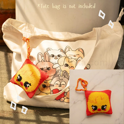 A cream canvas tote bag with a printed skzoo illustration on the front, shown alongside a small orange and yellow cross-stitch style skzoo bag hanging with a matching strap. The image includes text stating '*Tote bag is not included' and a small inset showing the pouch alone on a marble surface.