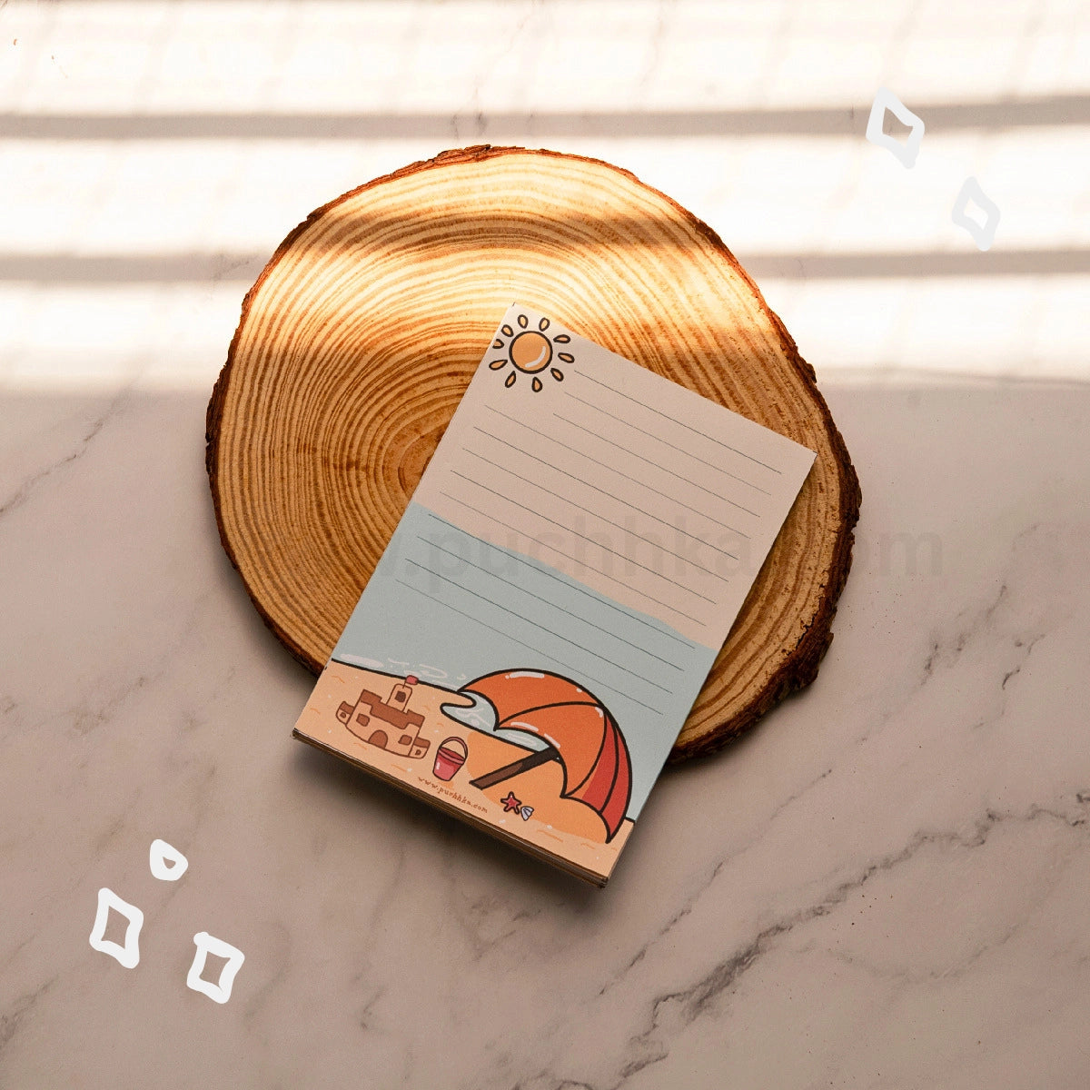 A small notepad with a beach illustration and lined writing area, placed on a round wood coaster over a light marble surface with soft window-shadow light.