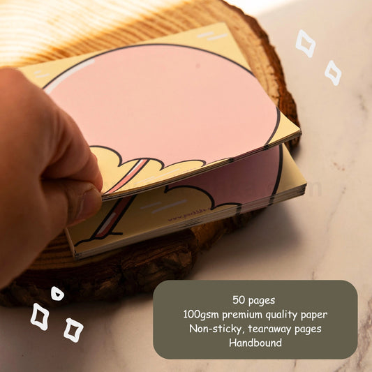 A hand holds a stack of tearaway memopad pages with a pink umbrella on a beach, resting on a rustic wood coaster over a light marble surface. A dark gray text box in the bottom right corner reads: '50 pages, 100gsm premium quality paper, Non-sticky, tearaway pages, Handbound'.