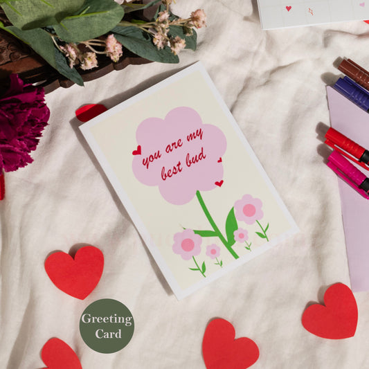 A square greeting card with a pink flower illustration and the text 'you are my best bud' lies on a cream fabric surface. The card is styled with red paper hearts, flowers, pens, and a green 'Greeting Card' badge.