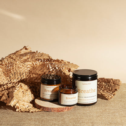 Three amber glass jars of Breathe candles with black lids and cream labels, styled on a burlap surface with decorative dried palm leaves against a warm beige background.