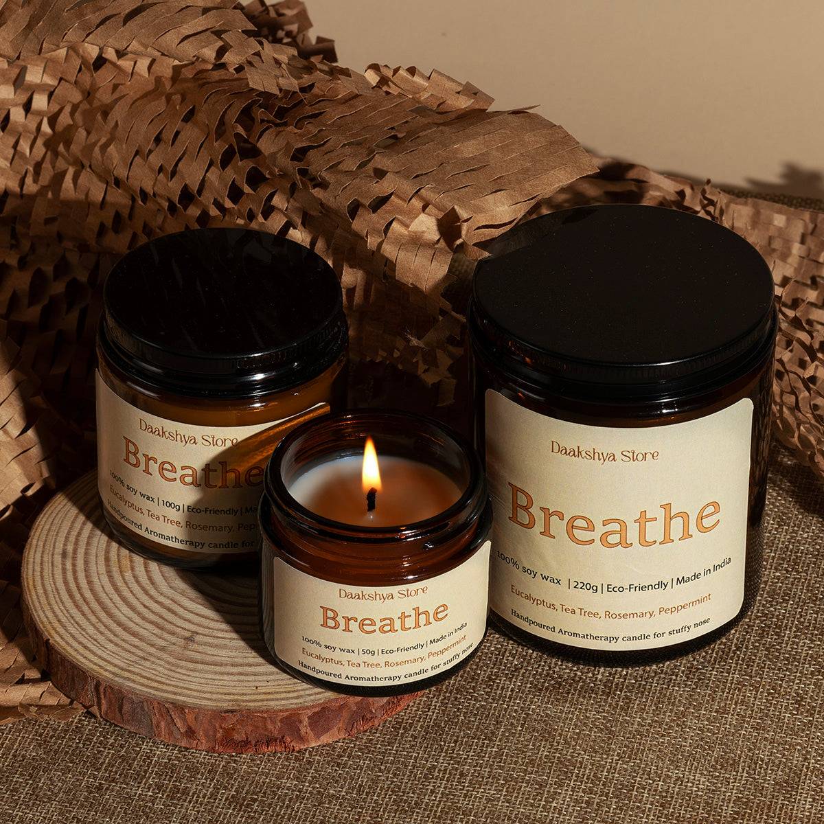 Three amber glass scented candles from Daakshya Store, including two jars and one lit candle, styled on a wooden slice with brown paper packaging in the background.