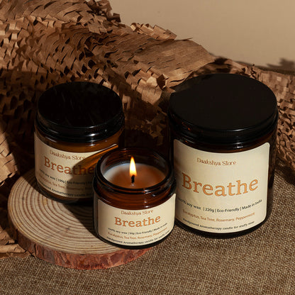 Three amber glass scented candles from Daakshya Store, including two jars and one lit candle, styled on a wooden slice with brown paper packaging in the background.