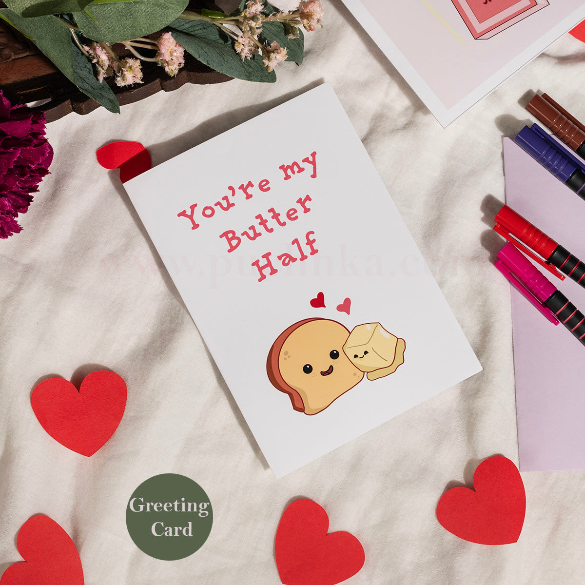 A white greeting card with the phrase 'You're my Butter Half' in pink text and a cute illustrated toast character hugging a small heart, styled on a cream fabric with red paper hearts, flowers, and stationery around it.