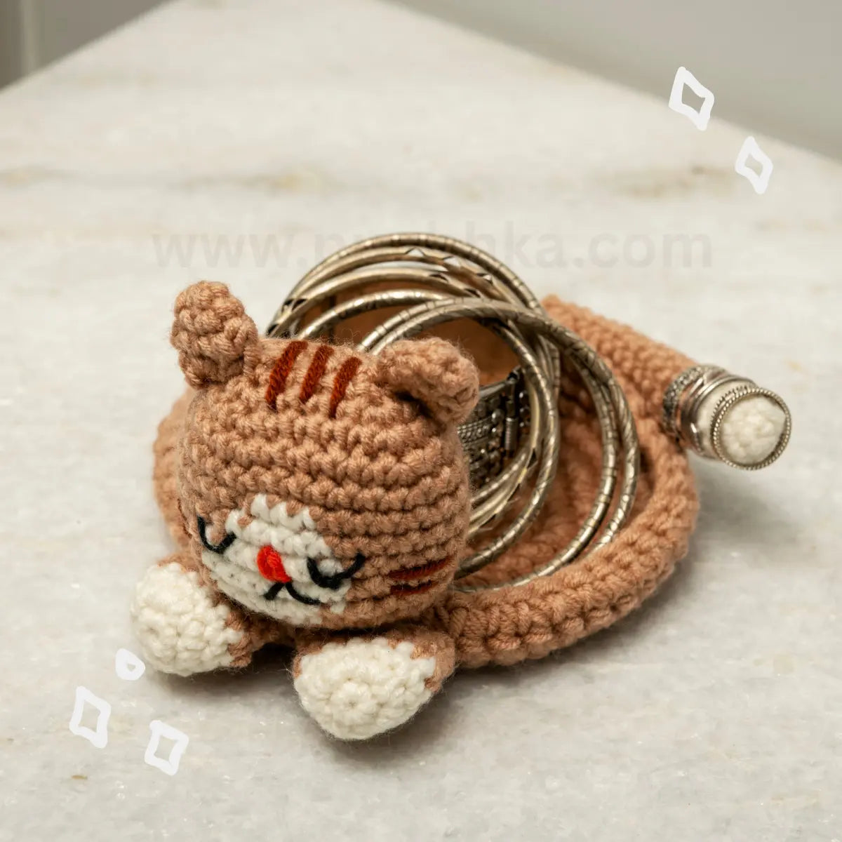 A crocheted cat-shaped tratorganizer with a metallic bangles resting on it. The cat is made from tan and brown yarn with white paws and a red nose, and the bangle is silver-toned.