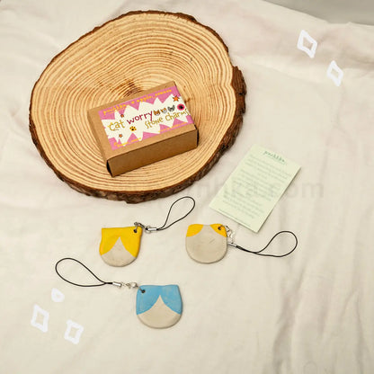 Three small cat-shaped phone strap charms in yellow, cream, and blue are arranged on a white fabric surface with a round wood slice, a small branded box, and a printed card.