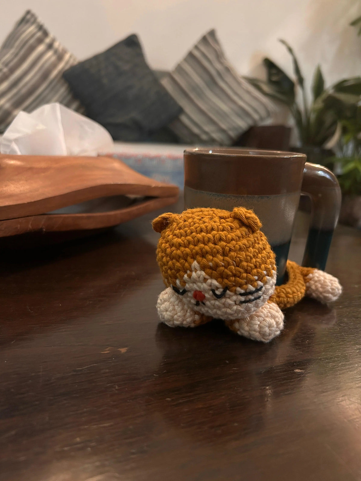A small crocheted cat-shaped tray in orange and cream sits on a dark wooden table and a mug resting on it. 