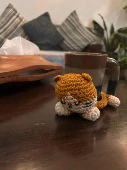A small crocheted cat-shaped tray in orange and cream sits on a dark wooden table and a mug resting on it. 