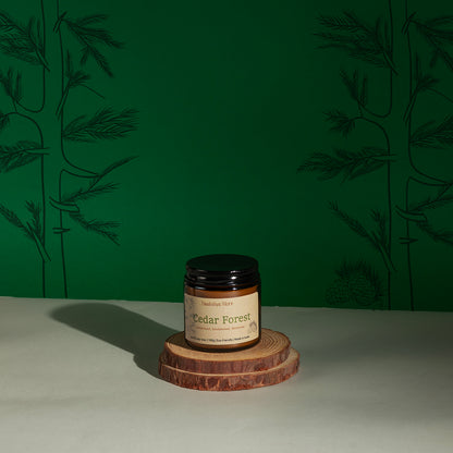 A jar of Cedar Forest scented candle by Daakshya Store (now Puchhka) on a stack of two wooden coaster, set against a dark green patterned background.