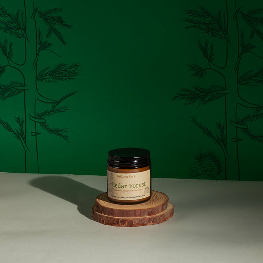 A jar of Cedar Forest scented candle by Daakshya Store (now Puchhka) on a stack of two wooden coaster, set against a dark green patterned background.