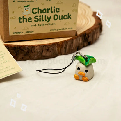 A small plush desk buddy charm shaped like a duck with a green leaf hat and orange feet, attached to a black cord, displayed on a white surface in front of its branded cardboard box on a wood background.