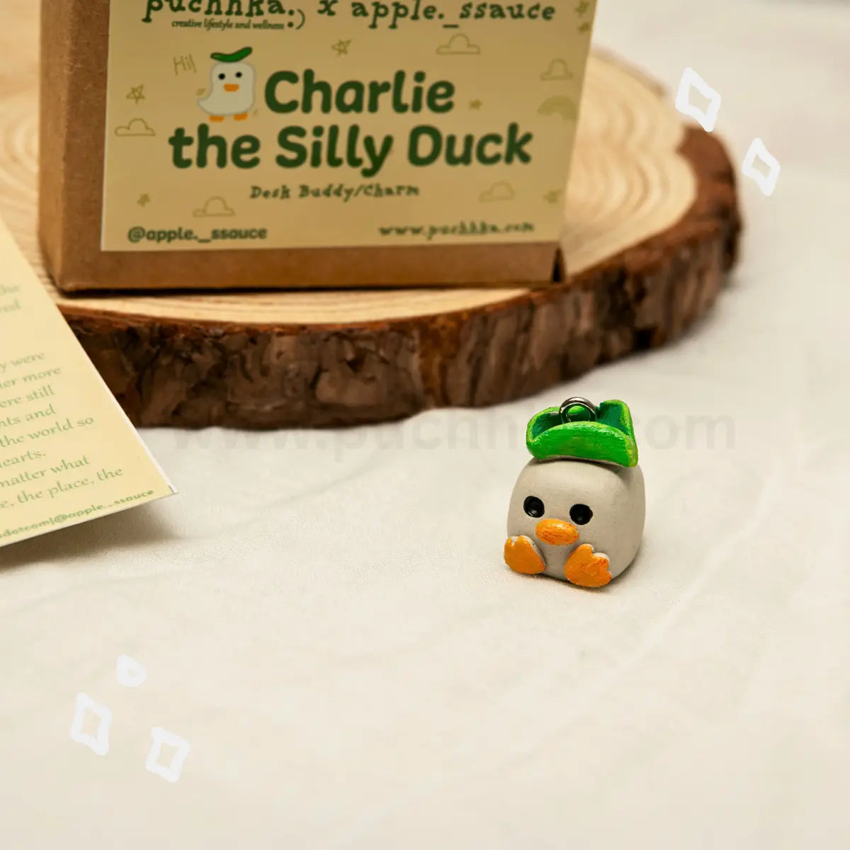 A small clay figurine of 'Charlie the Silly Duck' with a green hat and orange feet, displayed next to its branded packaging box on a rustic wood slice coaster.